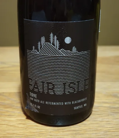 Tove bottle, Fair Isle: dark bottle with a black label showing a line drawing of a wooded island under a full moon above wavy water lines, text reading Fair Isle, Tove, oak-aged ale refermented with blackberries, 12.7 fl oz, Seattle, WA